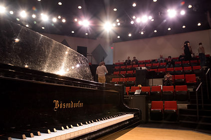 The front of the Bosendorfer piano to the left, with the red auditorium chairs in the background.