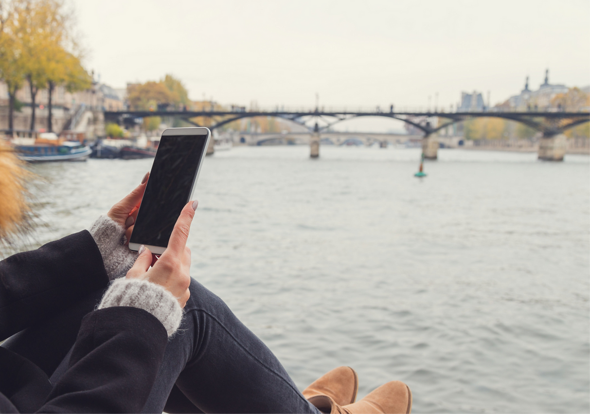 A photo looking over the shoulder of aperson sits on a riverside in a city looking at their phone screen.
