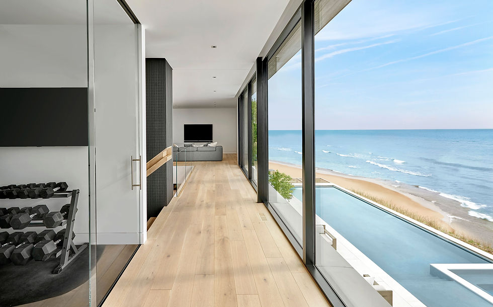 Light-filled corridor in a modern coastal home, with floor-to-ceiling glass overlooking the ocean and a lap pool, connecting a fitness area to a living space.