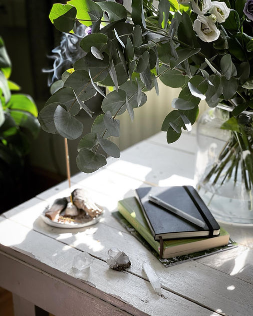 A stack of notebooks and books on white table, a lit incense is behind it. Greenery is also visible