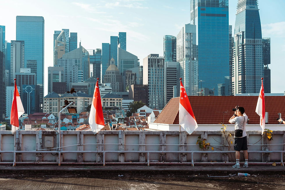 A person photographing a cityscape featuring skyscrapers, with Singapore flags in the foreground on a rooftop, under a clear blue sky.