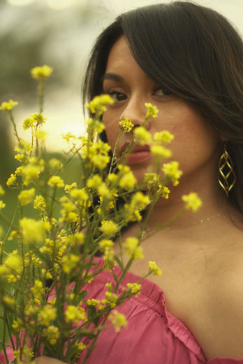 Woman in a flowing pink off-the-shoulder dress holding yellow wildflowers in a spring meadow surrounded by green trees and scattered wildflowers, captured during a natural light portrait session in Round Rock, Tx taken by an Austin, TX photographer- Ortega Photography.