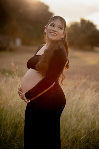 Maternity photoshoot at golden hour featuring a pregnant woman in a black dress holding wheat stems, smiling and looking over her shoulder in a natural outdoor setting with soft sunlight and tall grass in Austin, TX by Ortega Photography.