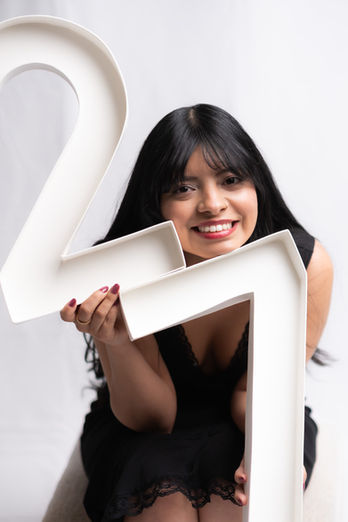 Smiling woman with long black hair in a black dress posing with large white 27 numbers in a soft, indoor setting, studio photography by Ortega Photography  in the Austin, Tx area.Smiling woman with long black hair in a white polka dotted dress posing with pink birthday cake in a soft, indoor setting, studio photography by Ortega Photography  in the Austin, Tx area.