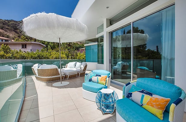 Turquoise outdoor chairs and colorful cushions beside rounded daybeds on a sunny hillside terrace.