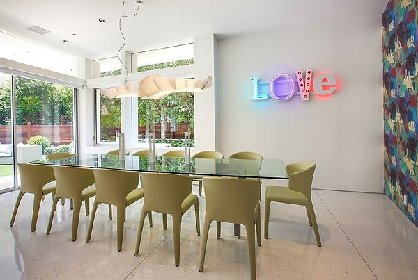 Glass dining table with pale green chairs beneath pendant lights, beside colorful wall covering and a neon LOVE sign.