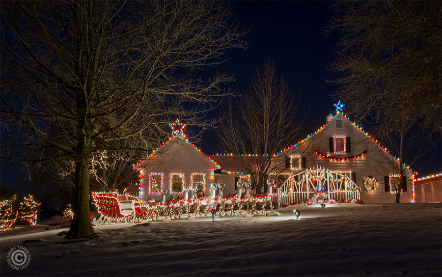 A sleigh and eight full sized reindeer in front of a house decorated with Christmas lights.