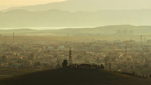 Hazy aerial view of a small city surrounded by rolling hills at sunrise, with soft golden light and power lines visible in the foreground