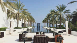 Luxury outdoor lounge with palm trees and a reflecting pool leading to the ocean under a clear blue sky.