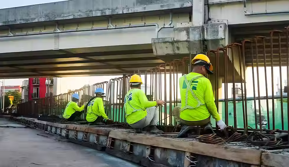 Hulagway sa mga construction worker nga nagsul-ob og safety helmet ug neon green nga uniporme samtang nagtrabaho sa mga puthaw sa ilawom sa tulay.