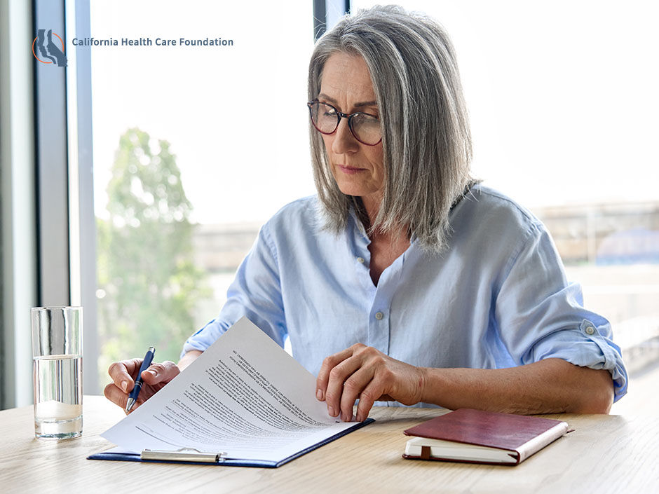 In a sunlit room, next to a large window looking out at an office building, a woman with silver hair looks through a legal document