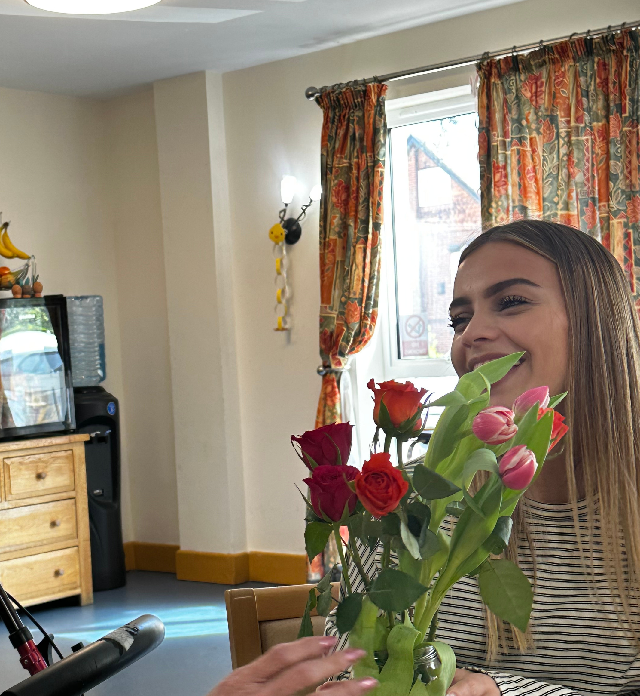 Elderly woman and smiling young woman share a vase of colorful roses and tulips at a table in a bright room.