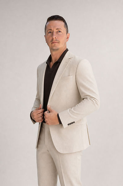Model Craig Mulgrew standing in a three-quarter pose wearing a beige suit and black polo shirt, holding the jacket lapels wit