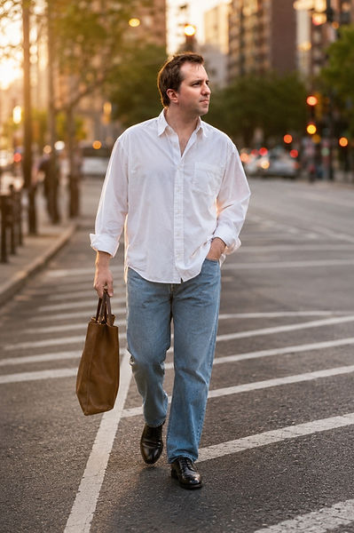 William in a white button-down and light-wash jeans carrying a brown tote while crossing a city street at sunset.