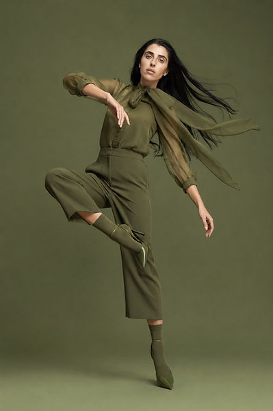 Ella Davis balancing on one leg in a dance-like pose, wearing a sheer olive pussy-bow blouse, trousers, socks and heels.