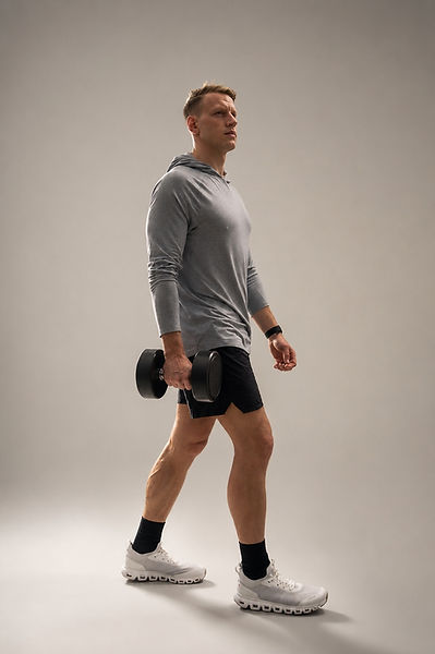 Fitness model Jesper Cedergren walking with a single dumbbell, grey hooded top, studio backdrop.
