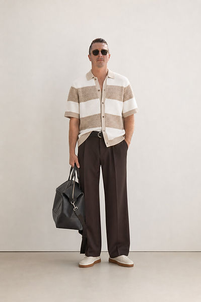 Model Craig Mulgrew standing in a beige and white striped knit polo, brown tailored trousers, and sunglasses, holding a black