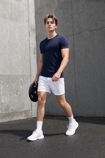 Ollie in navy t-shirt and white shorts walking outdoors, carrying a black weight plate against a concrete wall.
