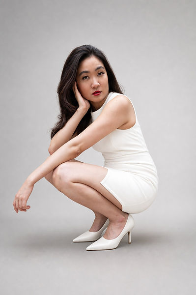 Caelyn in crouching in a white sleeveless dress and white heels, resting her head against her hand, against a gray background