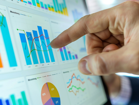 Hand pointing at colorful charts on a digital screen, showing various line and bar graphs. The mood is analytical and focused.