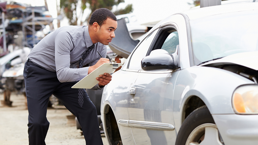 Man in a gray shirt inspects a damaged car with a clipboard at a salvage yard, focused expression, surrounded by stacked cars.