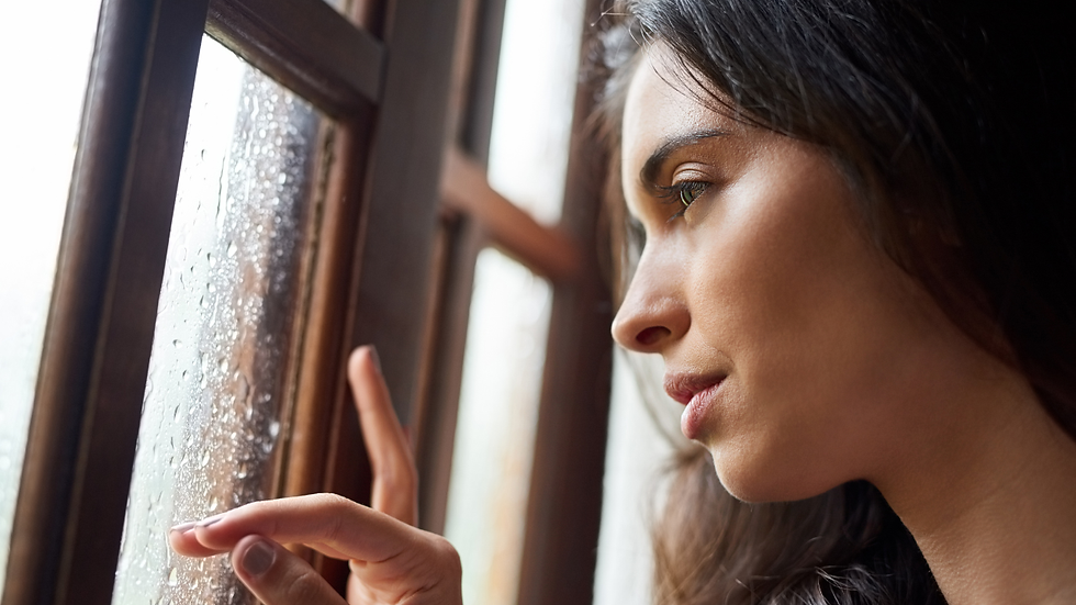 Woman gazes thoughtfully out a rain-streaked window, gently touching the glass. The wooden frame contrasts with the soft, serene mood.