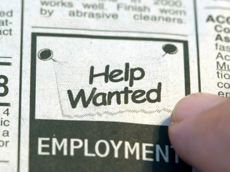 Close-up of a newspaper classified ad section with "Help Wanted" in bold. A finger points to the ad, suggesting job search activity.