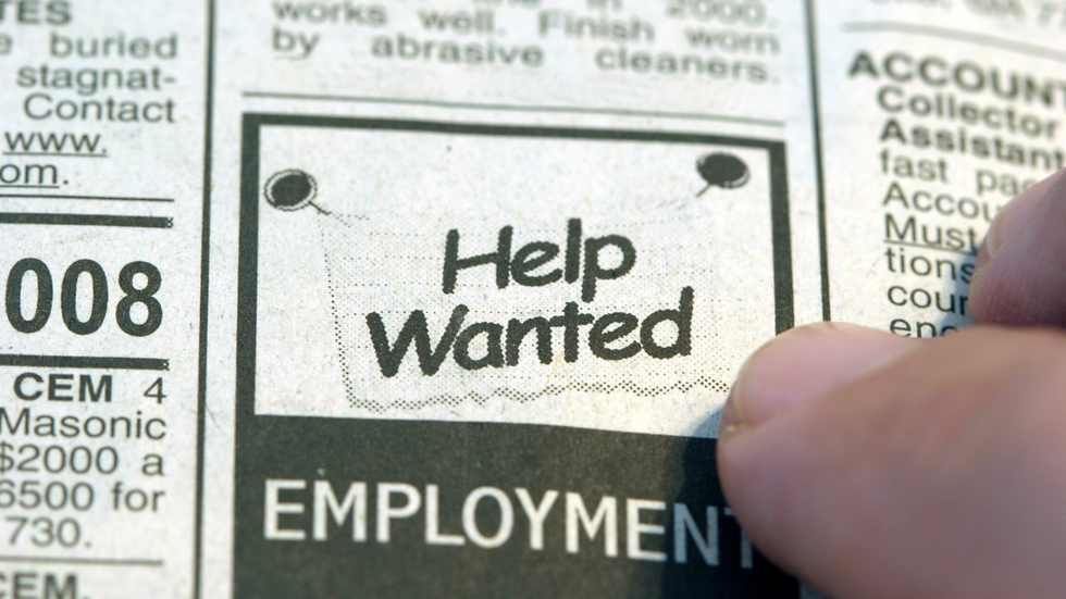 Close-up of a newspaper classified ad section with "Help Wanted" in bold. A finger points to the ad, suggesting job search activity.