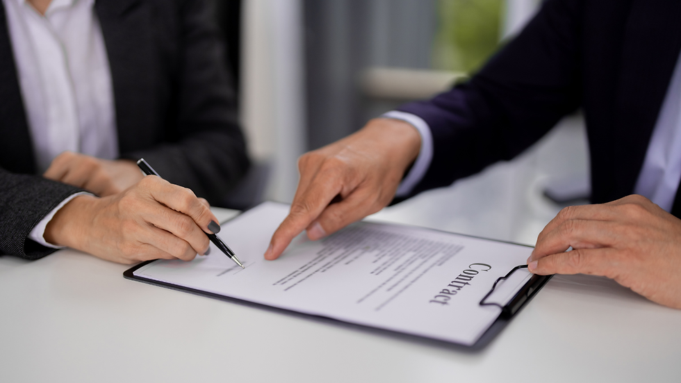 Hands sign a contract on a clipboard. One person points at the document. Both wear formal attire. The setting is a bright office.