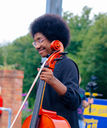 Millicent James, a Black woman with glasses and an afro, smiles to a person offscreen while holding a cello. In the background is the blue-and-yellow Ikon slow boat.