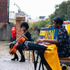 Jaz is sat outside, playing a keyboard. She is wearing a black jumpsuit, but has a bright yellow windbreaker draped over her lap. Millicent is sat near Jaz with her cello, looking towards Jaz's keyboard and pulling a surprised face with pursed lips.