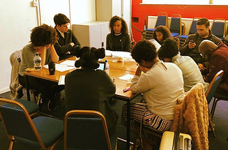 A script read-through, around two long tables pushed together. Jaz faces away from the camera, reading from a laptop in front of her, and listening as various actors read from their paper screenplays.