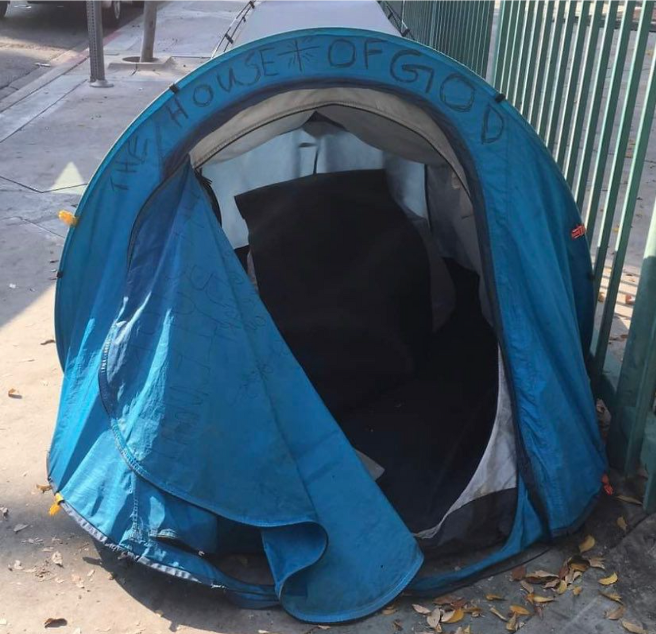 A tent sits on a pavement by a green gate. It is blue, round, and a little bit rough-looking, with it's door partially unzipped. Black marker has been scribbled on the tent's border. It reads, the house of God.