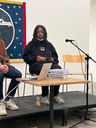 Jaz sits on a chair on a small platform at Ikon Gallery. A mic is on a stand and has been positioned for her to speak into. Jaz is mid-blink; she has a single-use mask tucked under her chin. Jacob Carter's arm and leg protrudes into the frame ever so slightly, as he is sat beside her. Jacob was in Ikon Youth Programme, and was in conversation with Jaz about the project they did.