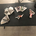 A bird's eye view of five paper designs on a table, like ornaments. One is a plane; two are fans; another, a bird; and the final one resembles a flower, with its lotus petals spread out.