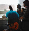Millicent plays her cello facing away from the camera, with only her cat-eye sunglasses, her blue afropuff hair, and the cello's scroll in view. Peaking over the top of her head are the side profiles of Jaz and the Word Life Corral, singing. Jaz is playing a keyboard.