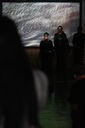 Due to rain, the second performance was moved into Centrala gallery. The lights are off, leaving hints of natural light and a blurry projection of Keith Piper's film to light the room. Over the shoulder of someone with dark hair, we see Two and a half of the Word Life Corral, with much of them hidden by the shadows.