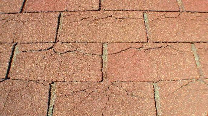Close-up of weathered red roof shingles with cracks and gaps, showing a rough, aged surface.
