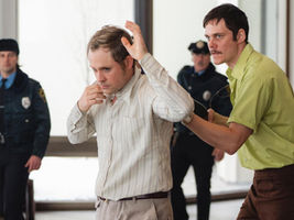 Un homme menace un autre homme avec un fusil dans le dos. Deux policiers regardent la scène.