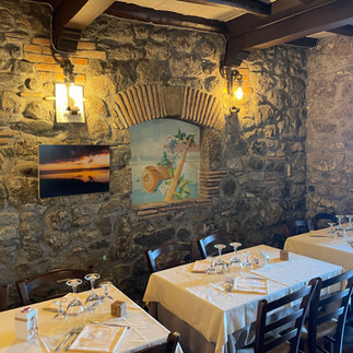 Ambiente accogliente con pareti in pietra, una nicchia ad arco con decorazioni e quadri appesi, tavoli apparecchiati con tovaglie bianche e sedie in legno, illuminazione d’atmosfera.