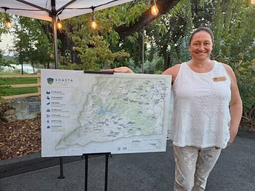 Woman smiling next to a detailed Shasta County map on an easel. She wears a white top, standing under a tent with string lights in a park.