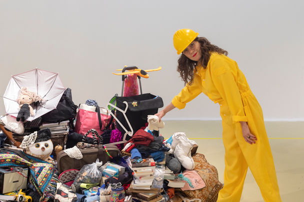 The Artist Dafna Talmon in yellow suit and helmet pointing to a pile of items. MIC - Made In China by Dafna Talmon