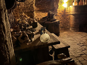 Ancient art of alchemy in Prague, Czech Republic. Dimly lit rustic cellar with stone walls, wooden shelves, pottery, and tools. A glowing lamp adds warmth. Quiet, historical ambiance.
