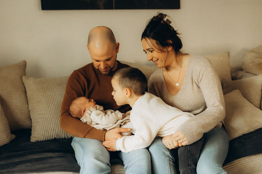 Eine Familie sitzt gemeinsam auf dem Sofa und bewundert liebevoll ihr Neugeborenes. Neugeborenenfotografie, die die innige Liebe zwischen Eltern und Geschwistern einfängt.  