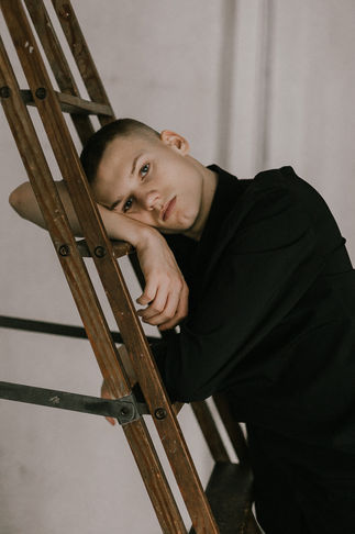 Halb-Portraitfoto des Schauspielers Caspar Hoffmann, der entspannt an einer Holzleiter lehnt und den Betrachter ansieht. 