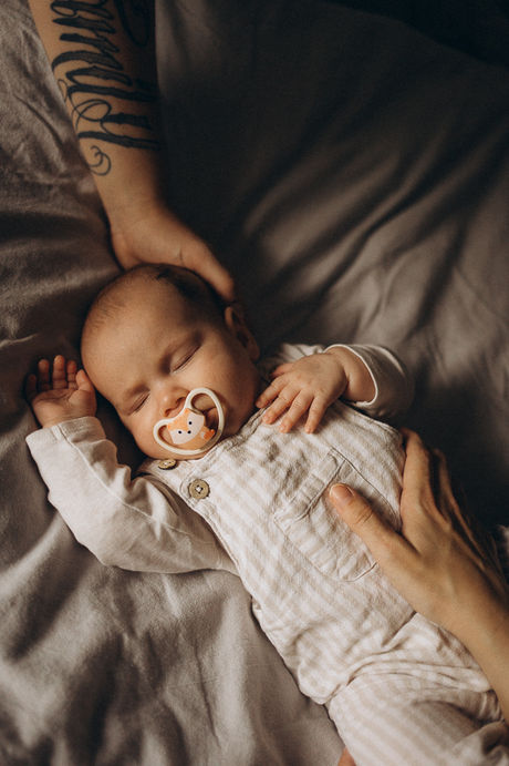Ein schlafendes Baby mit Schnuller liegt entspannt auf dem Bett, während die Hand des Vaters sanft seinen Kopf berührt und die Hand der Mutter sanft seinen Bauch. Babyfotografie, die Ruhe und Geborgenheit ausstrahlt. 