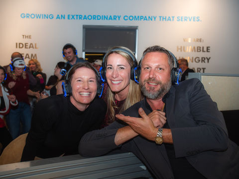 Three people wear headphones, smiling with company's text in background photography.