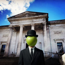 Man in suit and bowler hat with gree apple covering his face. Man stands in front of Stockport War Memorial Art Gallery