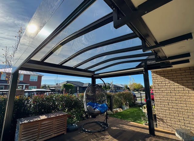 A clear, arched Transroof with black framing. There is an egg chair on the brown patio underneath the Transroof. 