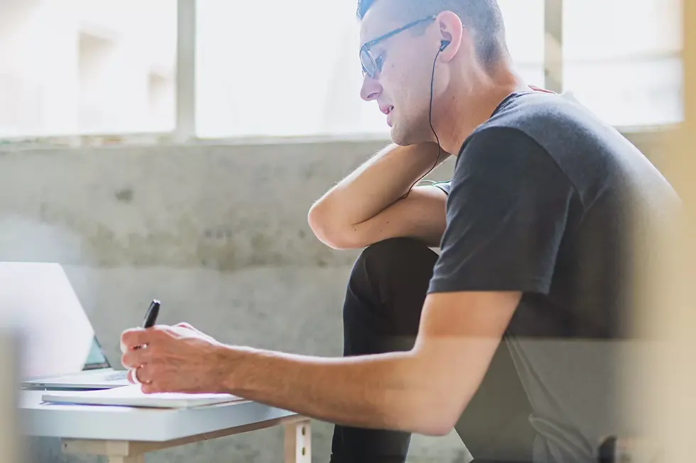 Person sitting at table, writing in book, with headphones on.